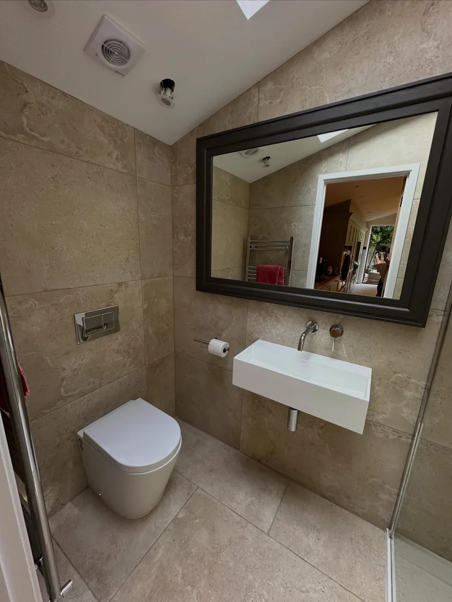A finished loft bathroom with a wall-hung basin, dark-framed mirror and beige stone tiles, fitted by Addisons